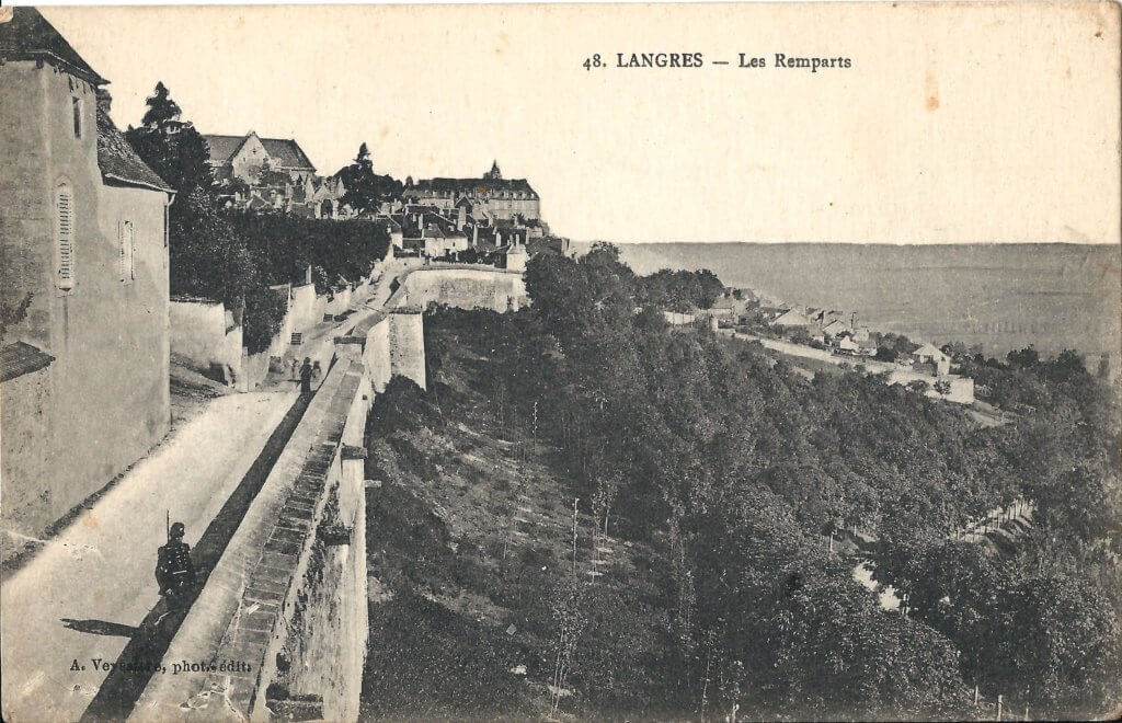 A postcard view of the old wall of the city of Langres, with a WWI era soldier patrolling, and wooded hillside below.